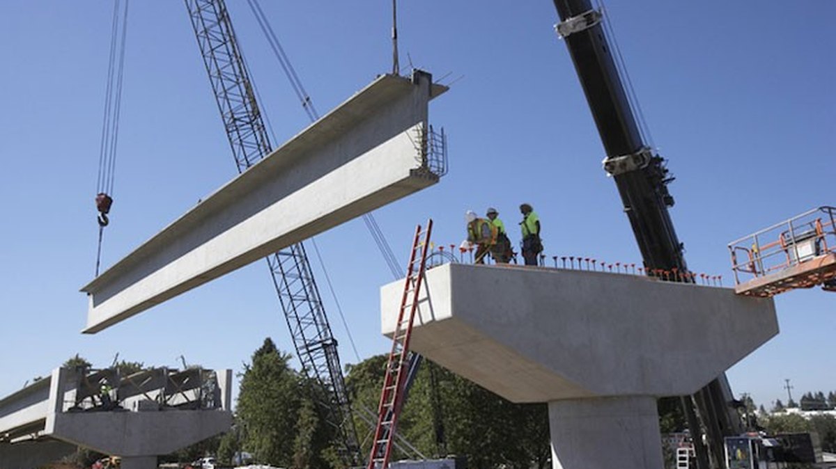 Cranes lowering a concrete bridge girder into place at a road construction site — OM Enterprise